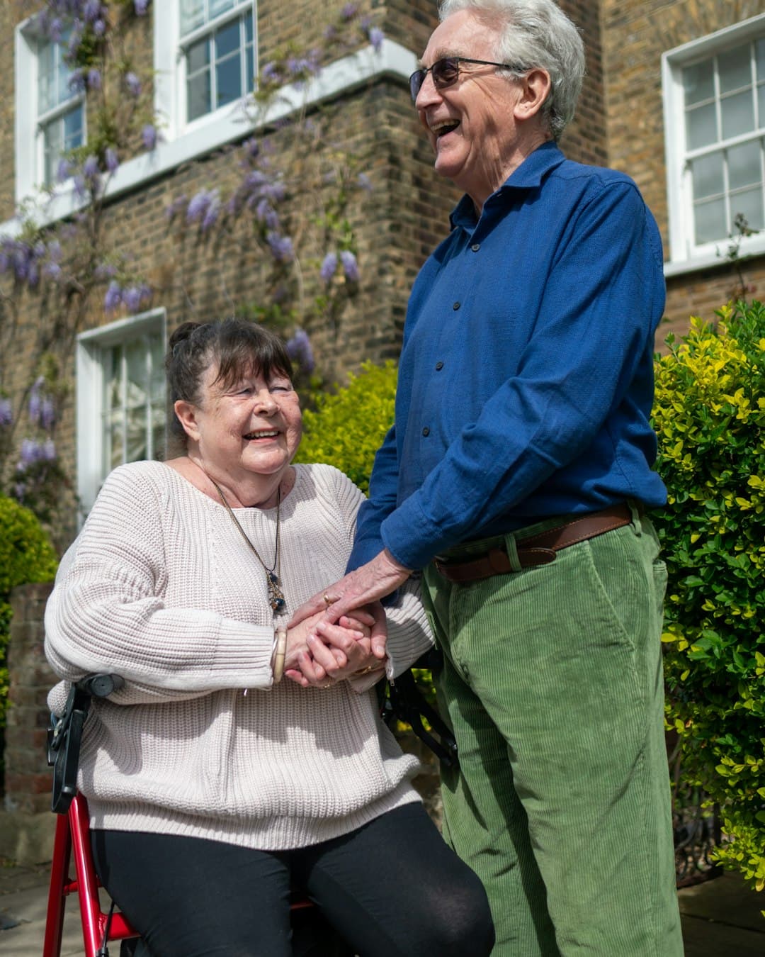 An older woman, seated, holding hands with a man standing beside her outside a house