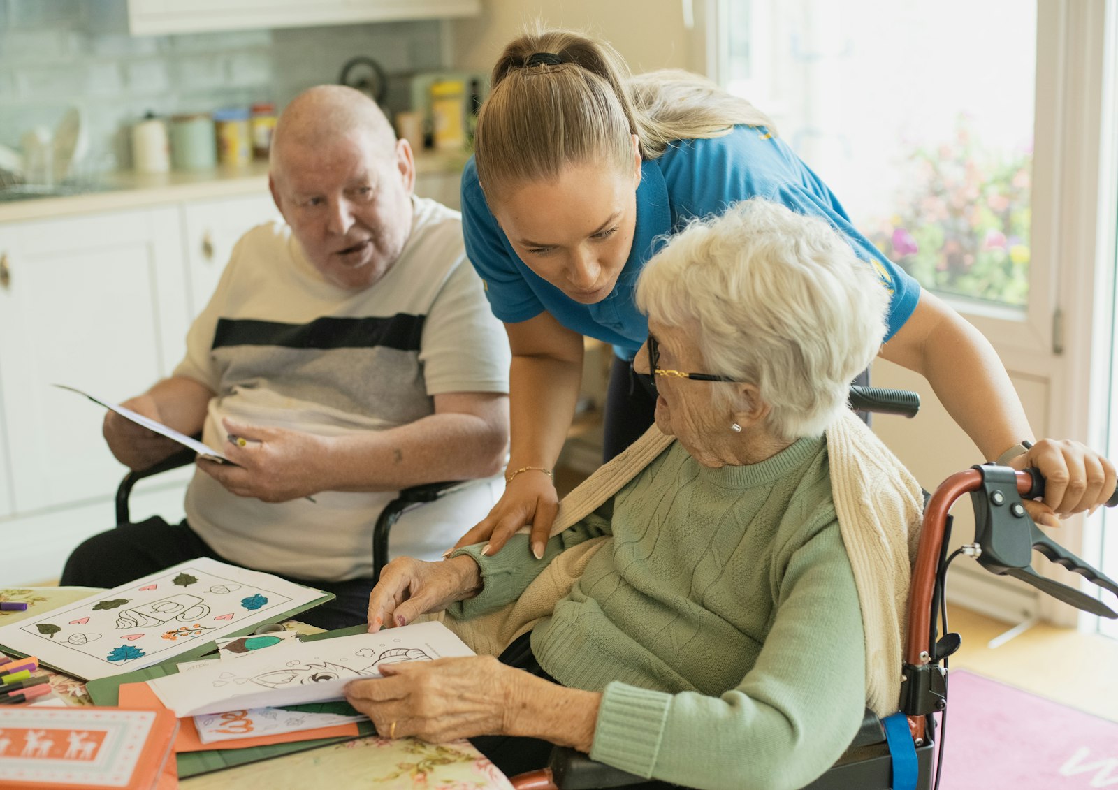A carer helping an older man and woman with a colouring activity at a table
